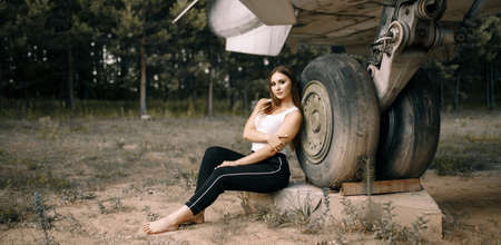 beautiful young brunette girl stands on the background of old military aircraft. Girl in a white shirt and black pants in nature. Military equipment. Portrait half-length. girl posingの写真素材