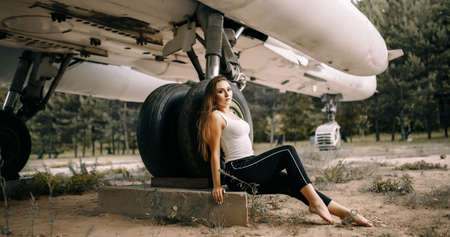 beautiful young brunette girl stands on the background of old military aircraft. Girl in a white shirt and black pants in nature. Military equipment. Portrait half-length. girl posingの写真素材