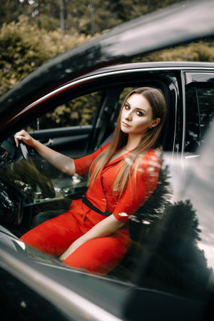 A beautiful young girl in a red overalls sits behind the wheel of a black car on an empty road in the forestの写真素材