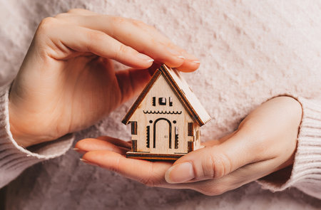 Woman holding a wooden house with her hands with the sun on a light pink background. Sweet homeの写真素材