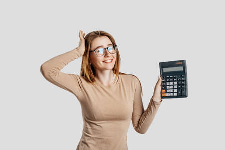 Beautiful young business woman wearing glasses holds calculator isolated on gray background. Achieving career wealth business accounting concept. Cover for advertisingの写真素材