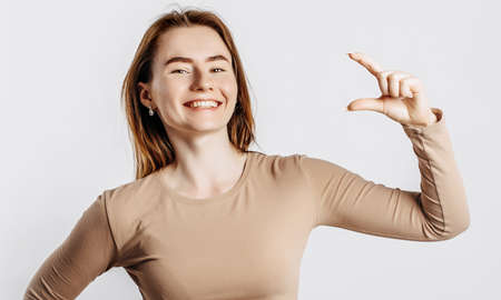 Young beautiful girl smiles and shows small size measure with her hand gesture on a white isolated background. A woman points to an idea, a place for advertising. Positive brunette in a beige jumper.の写真素材