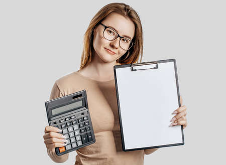 Beautiful young business woman wearing glasses holds calculator and clipboard with mock up blank space isolated on gray background. Achieving career wealth business concept. Cover for advertisingの写真素材