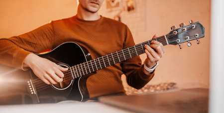 Man learning to play guitar with the help of online learning at home. Guy sitting at table with laptop and black guitarの写真素材