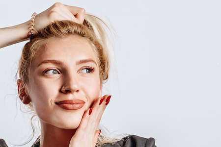 A beautiful blonde girl holds her hair and makes her own hairstyle with the emotion of happiness. Woman dreaming in black jacket and red top on gray isolated background for advertising. Nose piercing.の写真素材