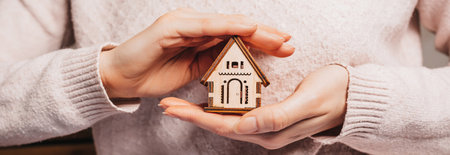 Woman holding a wooden house with her hands with the sun on a light pink background. Sweet homeの写真素材