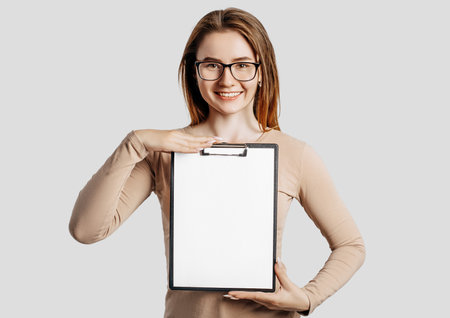 Beautiful young business woman wearing glasses holds a clipboard with mock up blank space isolated on gray background. Achieving career wealth business concept. Online learning coverの写真素材