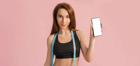 Fitness girl smiling and holding a phone with a mockup and posing hold measure tape in black sportswear on a pink background. Slim woman with a beautiful athletic body and tanned skinの写真素材