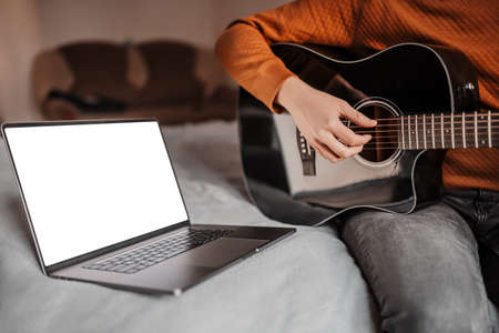 Man learning to play guitar with the help of online learning at home. Guy sitting on bed with laptop and black guitarの写真素材