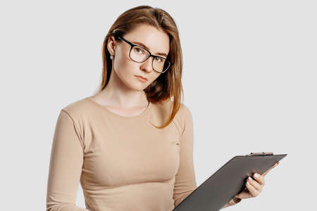 Beautiful young business woman wearing glasses isolated on gray background. Achieving career wealth business concept. Mockup copy space. Hold a clipboard with documentsの写真素材