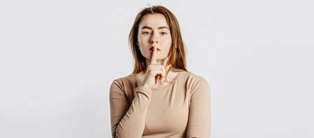 Shh gesture. Young beautiful serious girl holding a finger to her lips on a white isolated background. A woman asks to be silent, a place for advertising. Negative brunette in a beige jumper.の写真素材