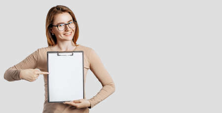 Beautiful young business woman wearing glasses holds a clipboard with mock up blank space isolated on gray background. Achieving career wealth business concept. Online learning coverの写真素材