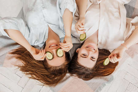 Two girls make homemade face and hair beauty masks. Cucumbers for the freshness of the skin around the eyes. Women take care of youthful skin. Girlfriends laugh at home lying on the floor on pillows.の写真素材