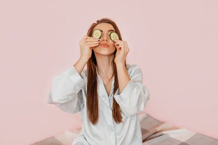 Girl make homemade face beauty masks. Cucumbers for the freshness of the skin around the eyes. Woman take care of youthful skin. Model laughing and having fun in spa on pink backgroundの写真素材