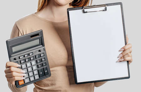 Young business woman holds calculator and clipboard with mock up blank space isolated on gray background. Achieving career wealth business concept. Cover for advertisingの写真素材