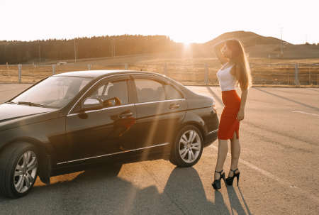 Beautiful young girl near a car in the evening in the sunset sun in an empty parking lotの写真素材