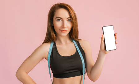 Fitness girl smiling and holding a phone with a mockup and posing hold measure tape in black sportswear on a pink background. Slim woman with a beautiful athletic body and tanned skinの写真素材