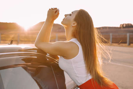 Beautiful young girl near a car in the evening in the sunset sun in an empty parking lotの写真素材