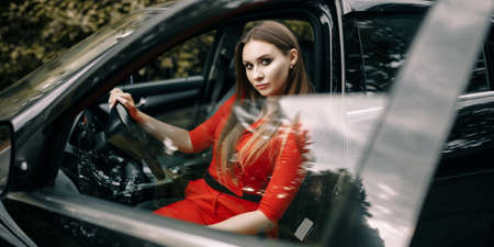 A beautiful young girl in a red overalls sits behind the wheel of a black car on an empty road in the forestの写真素材