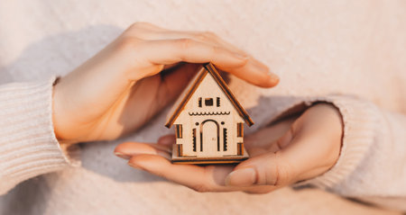 Woman holding a wooden house with her hands with the sun on a light pink background. Sweet homeの写真素材