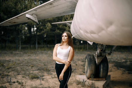 beautiful young brunette girl stands on the background of old military aircraft. Girl in a white shirt and black pants in nature. Military equipment. Portrait half-length. girl posingの写真素材