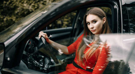 A beautiful young girl in a red overalls sits behind the wheel of a black car on an empty road in the forestの写真素材