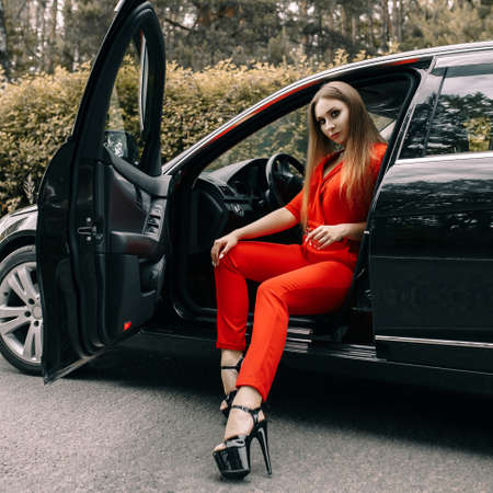 A beautiful young girl in a red overalls sits behind the wheel of a black car on an empty road in the forestの写真素材
