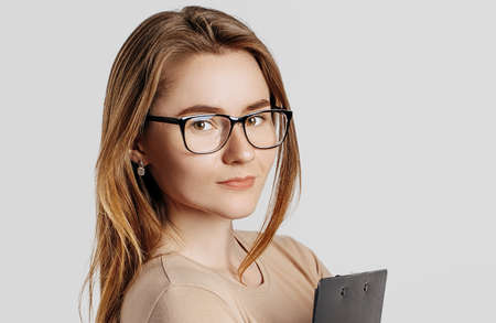 Beautiful young business woman wearing glasses isolated on gray background. Achieving career wealth business concept. Mockup copy space. Hold a clipboard with documentsの写真素材