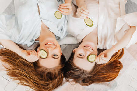 Two girls make homemade face and hair beauty masks. Cucumbers for the freshness of the skin around the eyes. Women take care of youthful skin. Girlfriends laugh at home lying on the floor on pillows.の写真素材