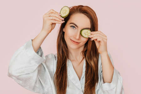 Girl make homemade face beauty masks. Cucumbers for the freshness of the skin around the eyes. Woman take care of youthful skin. Model laughing and having fun in spa on pink backgroundの写真素材