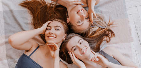 Women rest and have fun. Girlfriends laugh at home lying on the floor on pillows. Tree girls make homemade face and hair beauty masks. Women take care of youthful skin.の写真素材