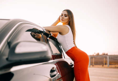 Beautiful young girl near a car in the evening in the sunset sun in an empty parking lotの写真素材
