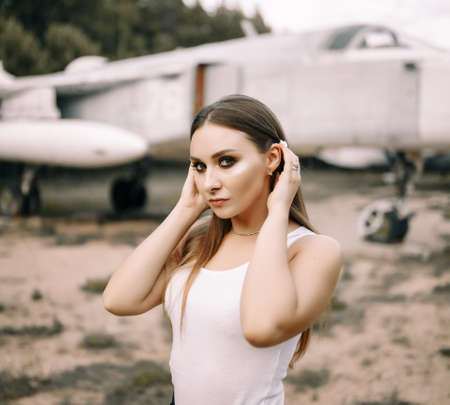 beautiful young brunette girl stands on the background of old military aircraft. Girl in a white shirt and black pants in nature. Military equipment. Portrait half-length. girl posingの写真素材