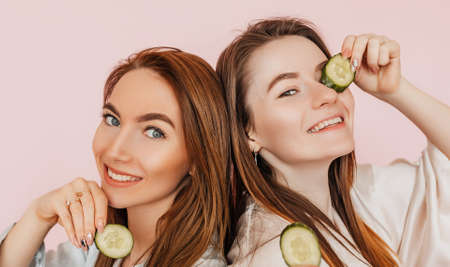 Two girls make homemade face beauty masks. Cucumbers for the freshness of the skin around the eyes. Women take care of youthful skin. Girlfriends laughing and having fun in spa on pink backgroundの写真素材