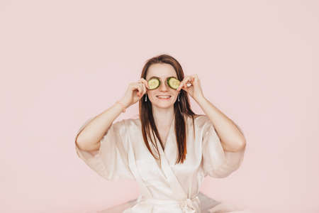 Girl make homemade face beauty masks. Cucumbers for the freshness of the skin around the eyes. Woman take care of youthful skin. Model laughing and having fun in spa on pink backgroundの写真素材