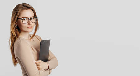 Beautiful young business woman wearing glasses isolated on gray background. Achieving career wealth business concept. Mockup copy space. Hold a clipboard with documentsの写真素材