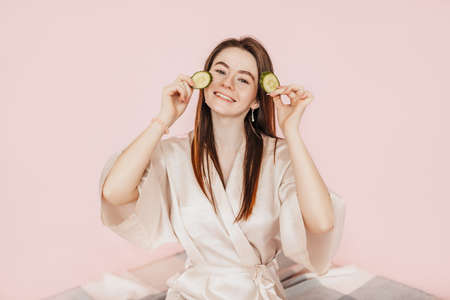 Girl make homemade face beauty masks. Cucumbers for the freshness of the skin around the eyes. Woman take care of youthful skin. Model laughing and having fun in spa on pink backgroundの写真素材