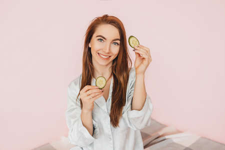 Girl make homemade face beauty masks. Cucumbers for the freshness of the skin around the eyes. Woman take care of youthful skin. Model laughing and having fun in spa on pink backgroundの写真素材