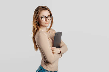 Beautiful young business woman wearing glasses isolated on gray background. Achieving career wealth business concept. Mockup copy space. Hold a clipboard with documentsの写真素材