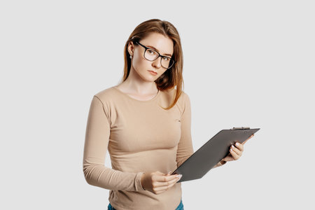 Beautiful young business woman wearing glasses isolated on gray background. Achieving career wealth business concept. Mockup copy space. Hold a clipboard with documentsの写真素材