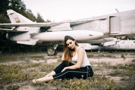 beautiful young brunette girl stands on the background of old military aircraft. Girl in a white shirt and black pants in nature. Military equipment. Portrait half-length. girl posingの写真素材