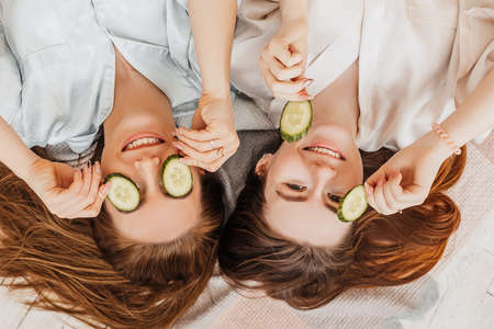 Two girls make homemade face and hair beauty masks. Cucumbers for the freshness of the skin around the eyes. Women take care of youthful skin. Girlfriends laugh at home lying on the floor on pillows.の写真素材