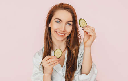 Girl make homemade face beauty masks. Cucumbers for the freshness of the skin around the eyes. Woman take care of youthful skin. Model laughing and having fun in spa on pink backgroundの写真素材