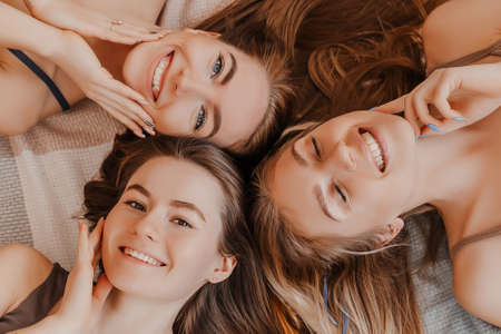 Women rest and have fun. Girlfriends laugh at home lying on the floor on pillows. Tree girls make homemade face and hair beauty masks. Women take care of youthful skin.の写真素材