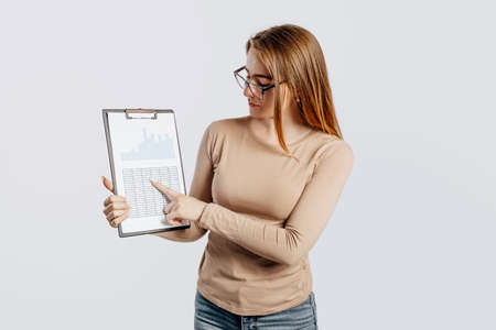 Beautiful young business woman wearing glasses isolated on gray background. Achieving career wealth business concept. Mockup copy space. Hold a clipboard with documents looking to the sideの写真素材
