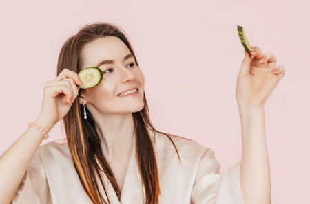 Girl make homemade face beauty masks. Cucumbers for the freshness of the skin around the eyes. Woman take care of youthful skin. Model laughing and having fun in spa on pink backgroundの写真素材
