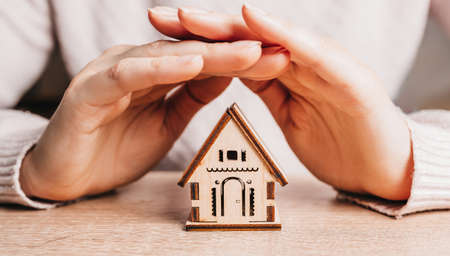 Woman holds and protects a wooden house with her hands with the sun on a light pink background. Sweet homeの写真素材