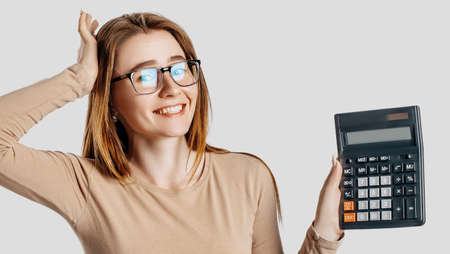 Beautiful young business woman wearing glasses holds calculator isolated on gray background. Achieving career wealth business accounting concept. Cover for advertisingの写真素材
