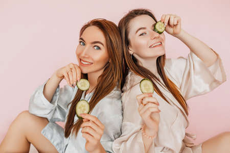 Two girls make homemade face beauty masks. Cucumbers for the freshness of the skin around the eyes. Women take care of youthful skin. Girlfriends laughing and having fun in spa on pink backgroundの写真素材