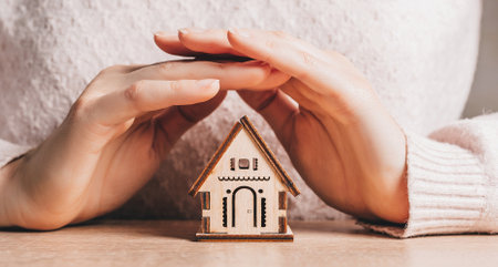 Woman holds and protects a wooden house with her hands with the sun on a light pink background. Sweet homeの写真素材